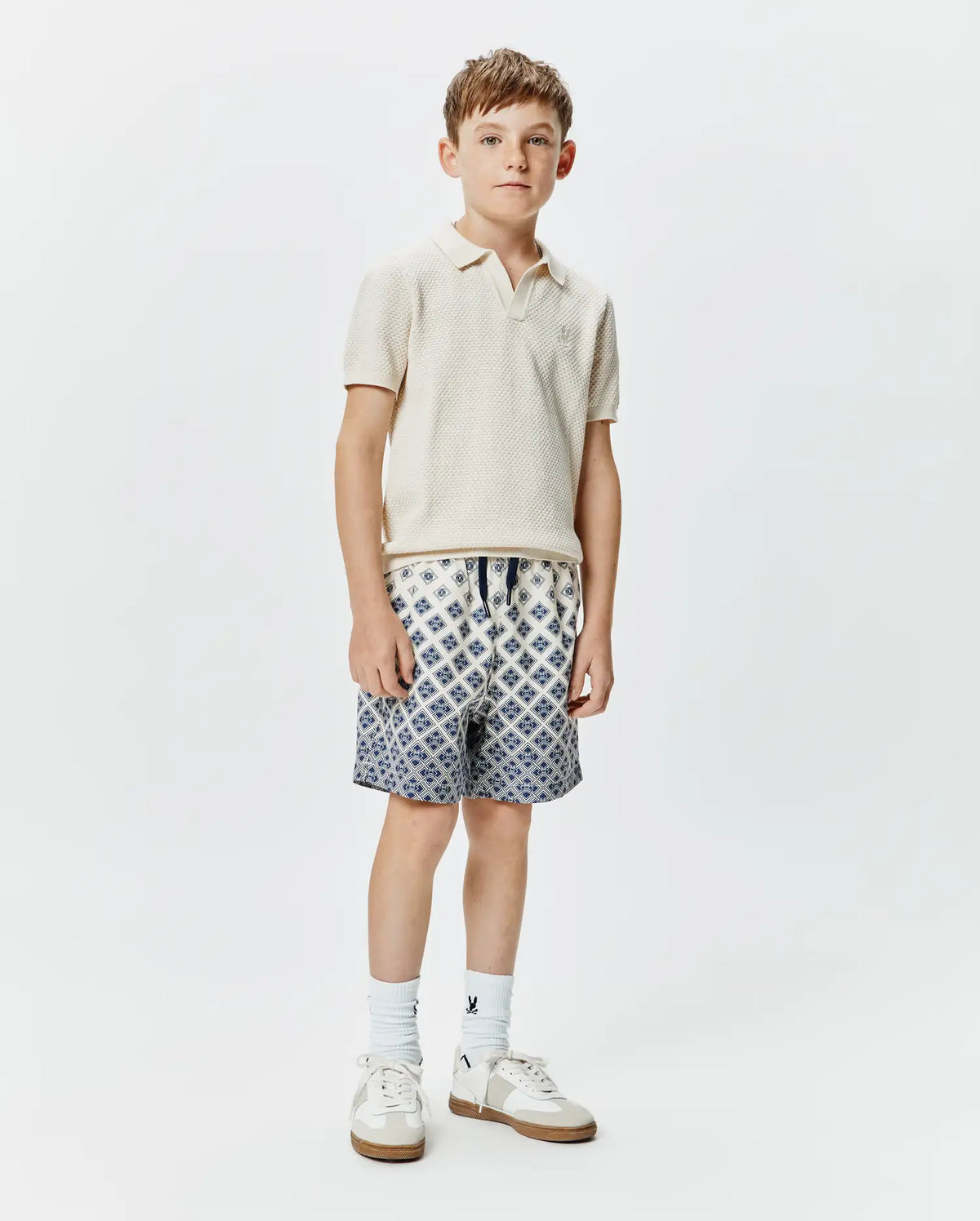 Classic fit cream pique knit polo with embroidered Psycho Bunny logo on chest, paired with Kids Tobias Swim Trunk featuring a white base and intricate blue geometric pattern, drawstring waist. (4)