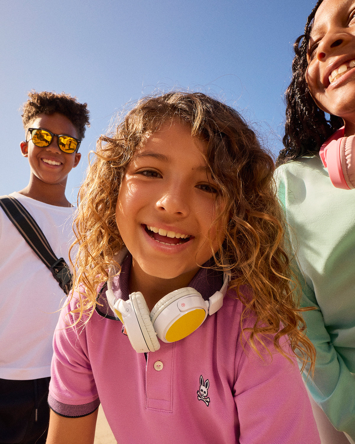Three smiling children, one wearing reflective sunglasses, one with white headphones around their neck, and another with pink headphones around their neck, enjoy a sunny day in Southport. Their brightly colored **Psycho Bunny KIDS SOUTHPORT PIQUE POLO SHIRT - B0K263B200** stands out against the clear blue sky.