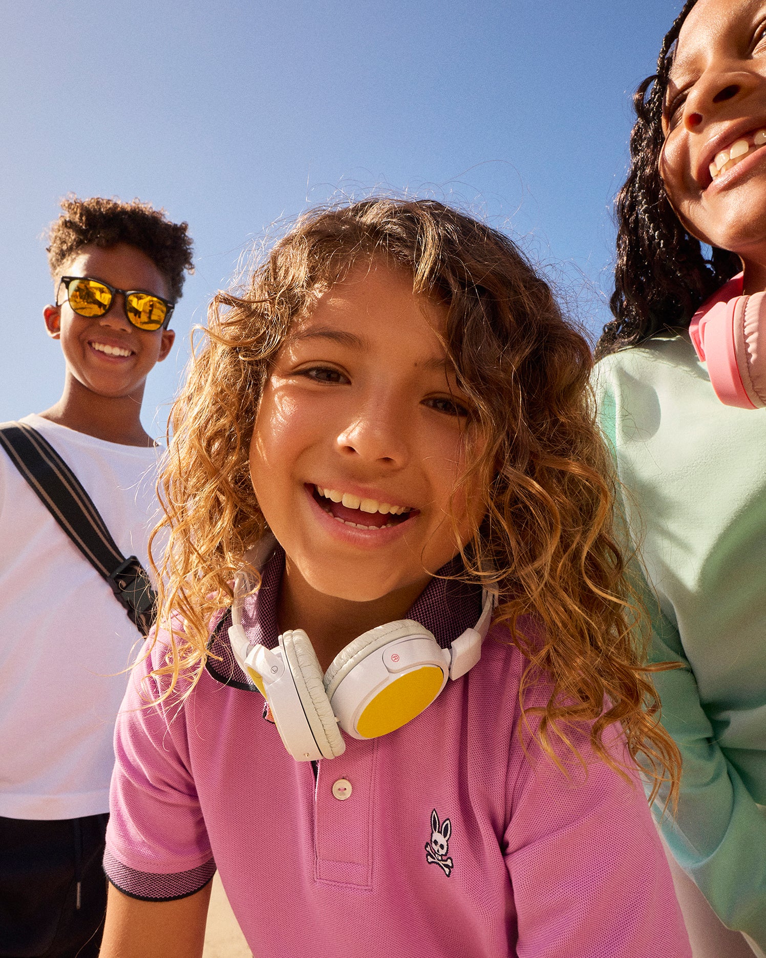 Three smiling children, one wearing reflective sunglasses, one with white headphones around their neck, and another with pink headphones around their neck, enjoy a sunny day in Southport. Their brightly colored **Psycho Bunny KIDS SOUTHPORT PIQUE POLO SHIRT - B0K263B200** stands out against the clear blue sky.