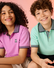 Two children smile and pose against a pink background. The child on the left wears a purple KIDS SOUTHPORT PIQUE POLO SHIRT - B0K263B200 by Psycho Bunny with a small rabbit logo, while the child on the right sports a light green polo. Both Pima cotton shirts feature dark contrasting collars and cuffs.