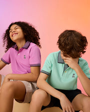 Two kids sit against a peach to orange gradient background. The child on the left, with curly hair, wears a pink KIDS SOUTHPORT PIQUE POLO SHIRT by Psycho Bunny and smiles. The child on the right, with wavy hair, dons a mint green KIDS SOUTHPORT PIQUE POLO SHIRT by Psycho Bunny and playfully covers their face with one hand.