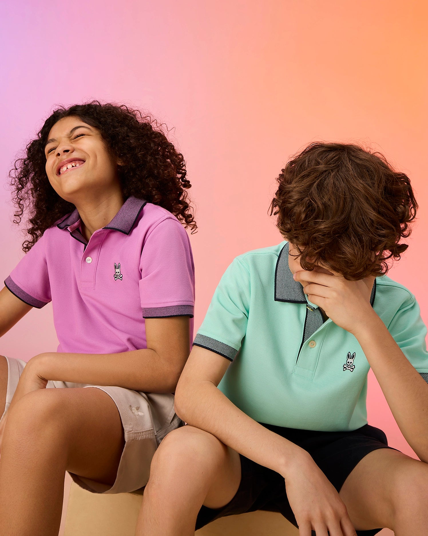 Two kids sit against a peach to orange gradient background. The child on the left, with curly hair, wears a pink KIDS SOUTHPORT PIQUE POLO SHIRT by Psycho Bunny and smiles. The child on the right, with wavy hair, dons a mint green KIDS SOUTHPORT PIQUE POLO SHIRT by Psycho Bunny and playfully covers their face with one hand.
