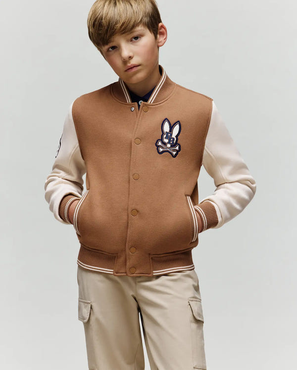 A young boy with light brown hair wears the Psycho Bunny KIDS WADE KNIT BOMBER (KO1100001) in tan and cream, featuring a bunny patch on the chest, with beige cargo pants. He stands against a plain light background, hands in pockets.