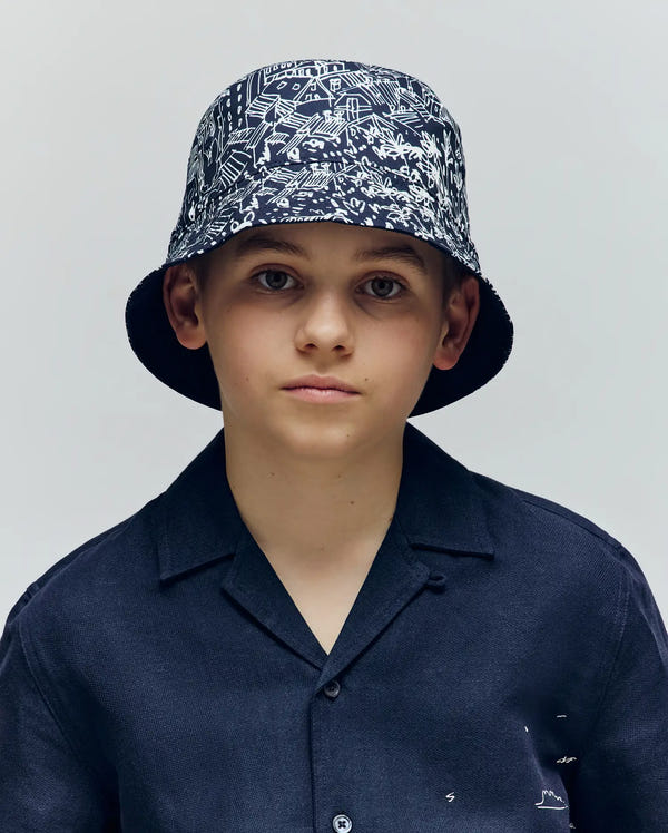 A boy in a dark, short-sleeved button-up shirt and the Psycho Bunny KIDS NAXOS REVERSIBLE BUCKET HAT (KO1500001) with a black-and-white pattern looks at the camera against a plain light background.