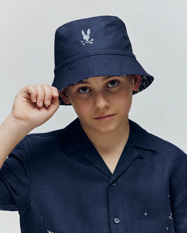 A young boy wears the Psycho Bunny KIDS NAXOS REVERSIBLE BUCKET HAT (KO1500001), featuring a white bunny and crossbones logo, as he looks at the camera and touches the quilted brim of his vibrant hat.
