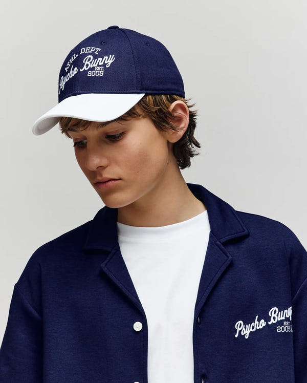 A young person wears a navy KIDS MATTEO BASEBALL CAP by Psycho Bunny, featuring a varsity style, white brim, and embroidered Psycho Bunny wordmark in white. Both cap and cotton twill jacket match. The background is plain and light.