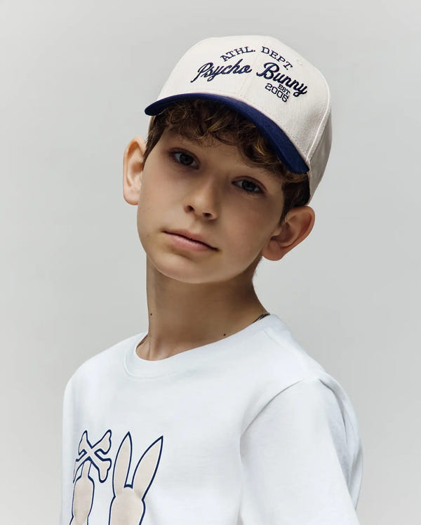 A young boy with light skin and curly brown hair wears the Psycho Bunny KIDS MATTEO BASEBALL CAP (KO1500002) in white and navy with an adjustable strap, looking at the camera with a neutral expression against a plain background.