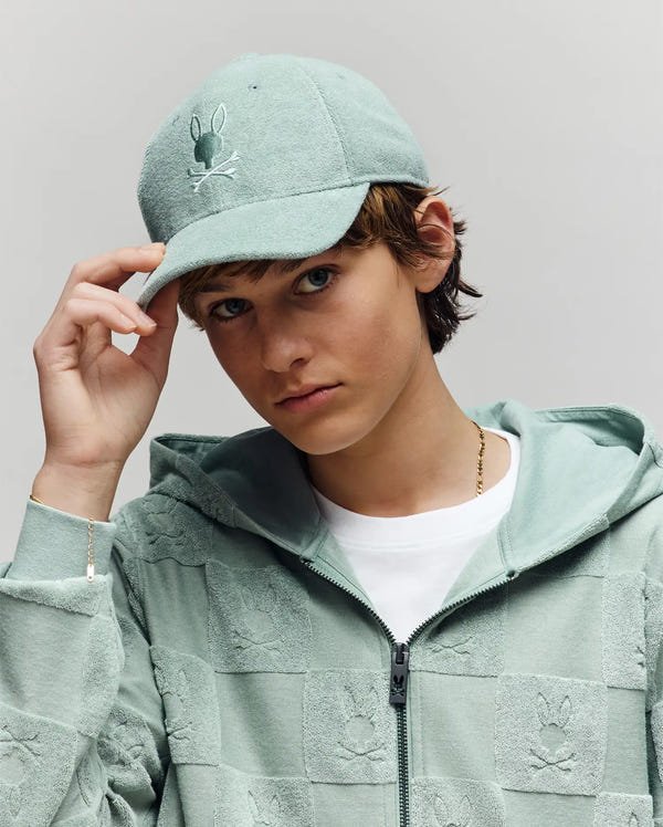 A young person in a light green Psycho Bunny KIDS KELVIN TOWEL TERRY BASEBALL CAP (KO1500007) with an embroidered Bunny logo and matching zip-up hoodie poses against a plain light gray background, holding the cap's brim.