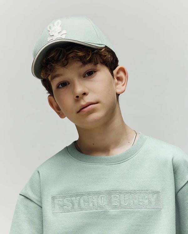 A young boy in a mint green premium cotton sweatshirt wears the Psycho Bunny KIDS DEREK BASEBALL CAP (KO1500008) with the classic logo, looking at the camera against a plain light gray background.