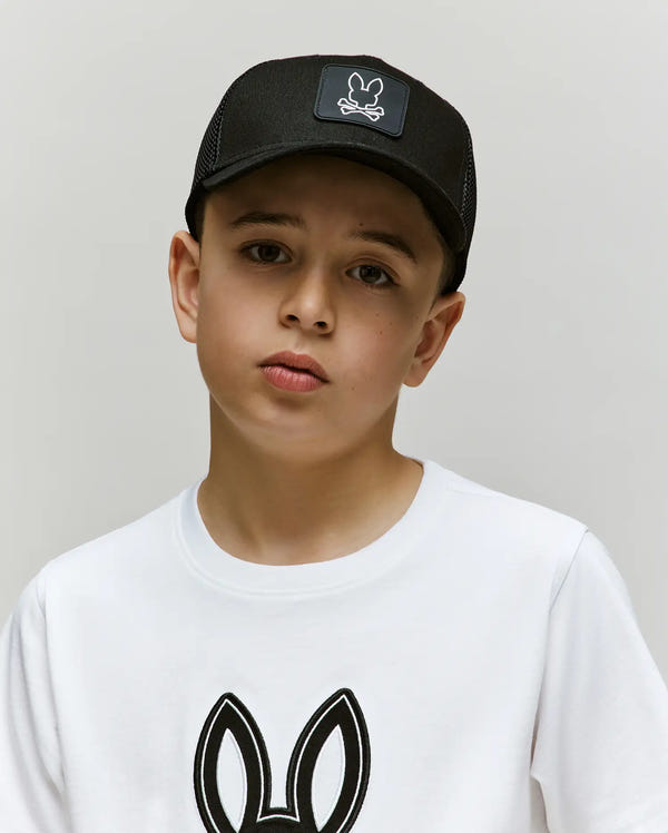A young boy wears the Psycho Bunny KIDS DARIO TRUCKER CAP (KO1500009) with a bunny logo and adjustable snap-back, paired with a white t-shirt featuring a matching bunny design, standing against a light gray background.