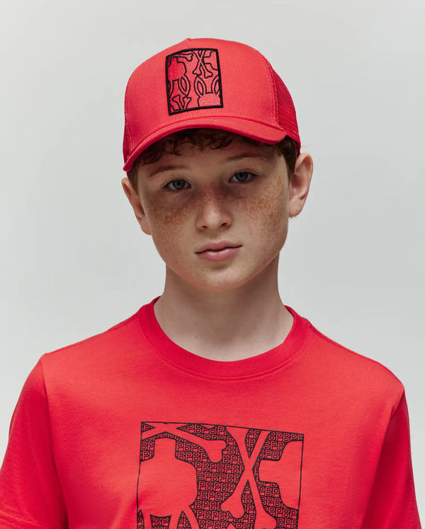 A young boy with light skin, brown hair, and freckles wears a Psycho Bunny KIDS GLENN TRUCKER CAP (KO1500012) in red with snapback fastening and matching T-shirt, both featuring a black rectangular graphic design, against a plain background.