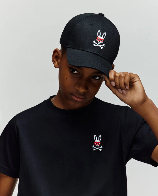 A young person in a black 100% cotton Mischief Bunny tee and Psycho Bunny KIDS MISCHIEF BASEBALL CAP (KO1500013), both with the white and red skull-and-crossbones bunny logo, stands against a plain light background, tilting the cap down.