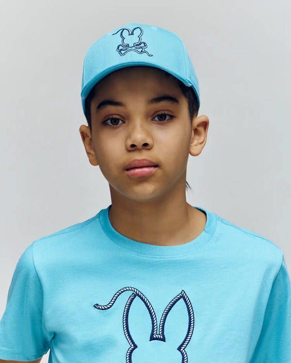 A young person wears the Psycho Bunny KIDS CYRUS BASEBALL CAP (KO1500014) and a matching t-shirt, both featuring a rabbit head design, standing against a plain, light background.
