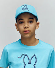 A young person wears the Psycho Bunny KIDS CYRUS BASEBALL CAP (KO1500014) and a matching t-shirt, both featuring a rabbit head design, standing against a plain, light background.