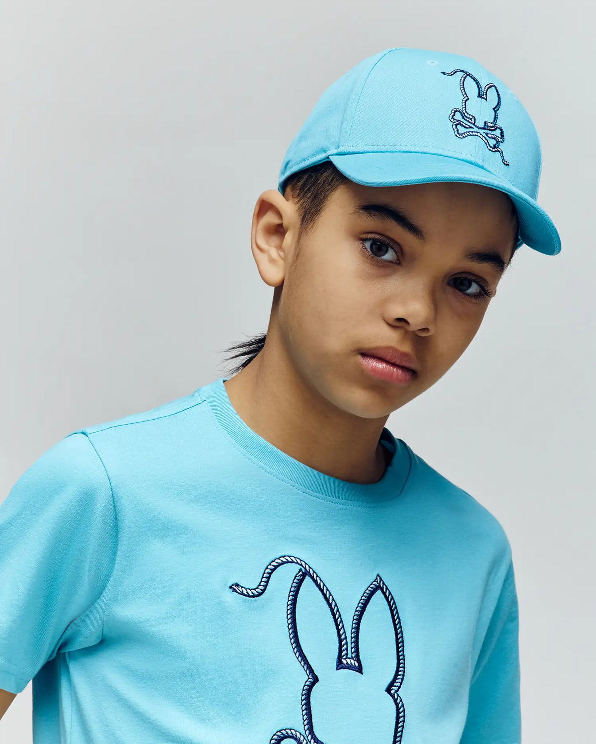 A young boy wears the Psycho Bunny KIDS CYRUS BASEBALL CAP (KO1500014) and a matching T-shirt with a bunny and rope graphic, standing against a plain background with a neutral expression.