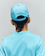 A child with braided hair is seen from behind, wearing a Psycho Bunny KIDS CYRUS BASEBALL CAP - KO1500014 in light blue cotton twill with "PG" embroidery and a matching T-shirt. The background is plain and light-colored.