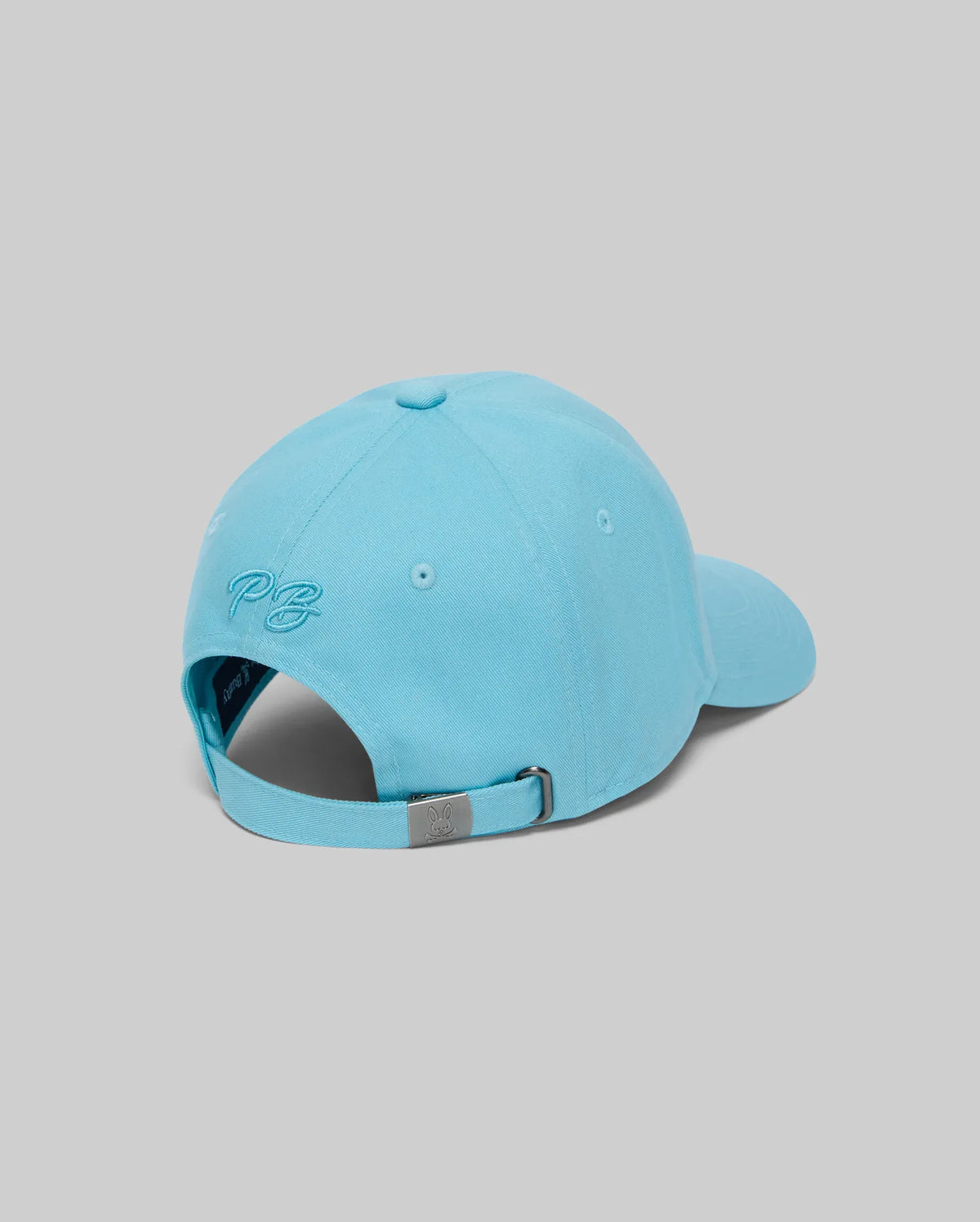 The KIDS CYRUS BASEBALL CAP by Psycho Bunny (KO1500014) is a light blue cotton twill cap, viewed from behind with "P.B." embroidered above an adjustable strap and metal clasp, adding coastal charm against a grey background.