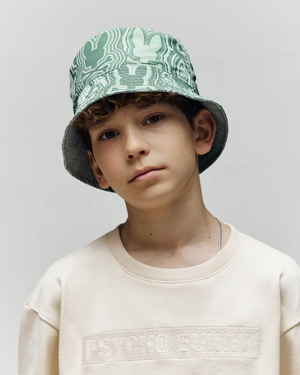 A young boy with curly hair wears the Psycho Bunny KIDS CHUCK REVERSIBLE BUCKET HAT (KO1500015), styled with a cream "PSYCHO BUNNY" sweatshirt, standing against a plain light background.