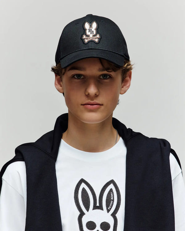 A young person wearing a Psycho Bunny KIDS WADE BASEBALL CAP (KO1500016) in black and a white tee, both featuring the embroidered Bunny logo in soft cotton twill, with a black jacket over their shoulders, stands against a light gray background.