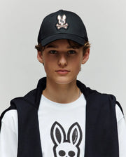 A young person wearing a Psycho Bunny KIDS WADE BASEBALL CAP (KO1500016) in black and a white tee, both featuring the embroidered Bunny logo in soft cotton twill, with a black jacket over their shoulders, stands against a light gray background.