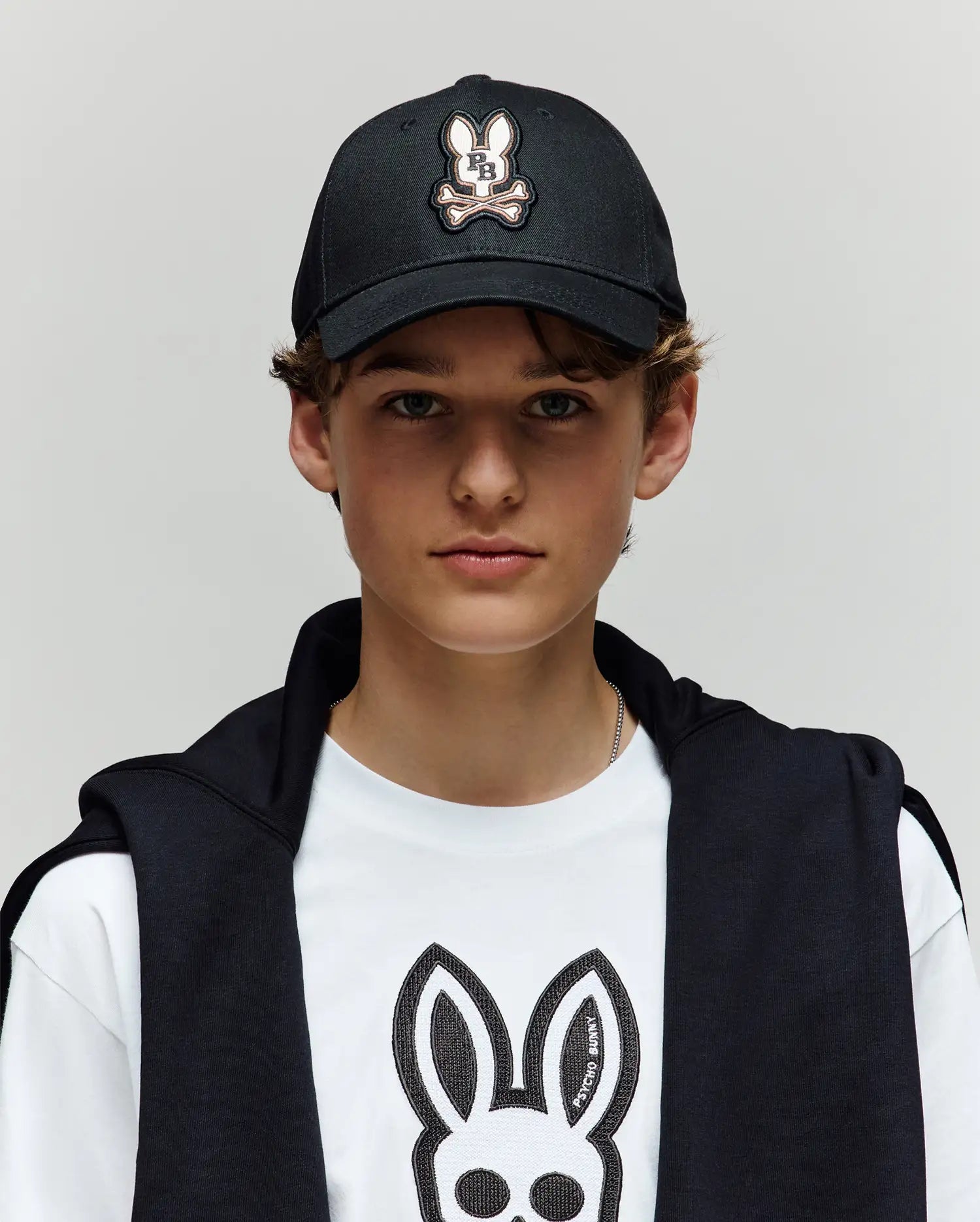 A young person wearing a Psycho Bunny KIDS WADE BASEBALL CAP (KO1500016) in black and a white tee, both featuring the embroidered Bunny logo in soft cotton twill, with a black jacket over their shoulders, stands against a light gray background.
