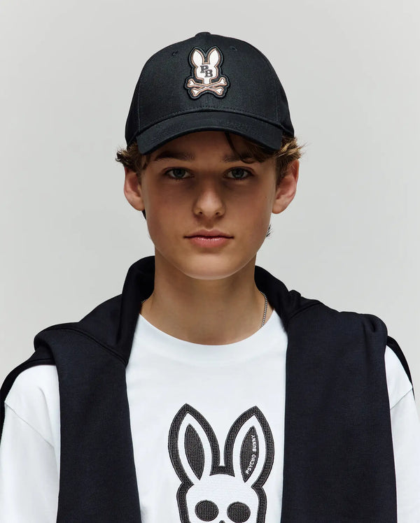 A young person wearing a Psycho Bunny KIDS WADE BASEBALL CAP (KO1500016) in black and a white tee, both featuring the embroidered Bunny logo in soft cotton twill, with a black jacket over their shoulders, stands against a light gray background.