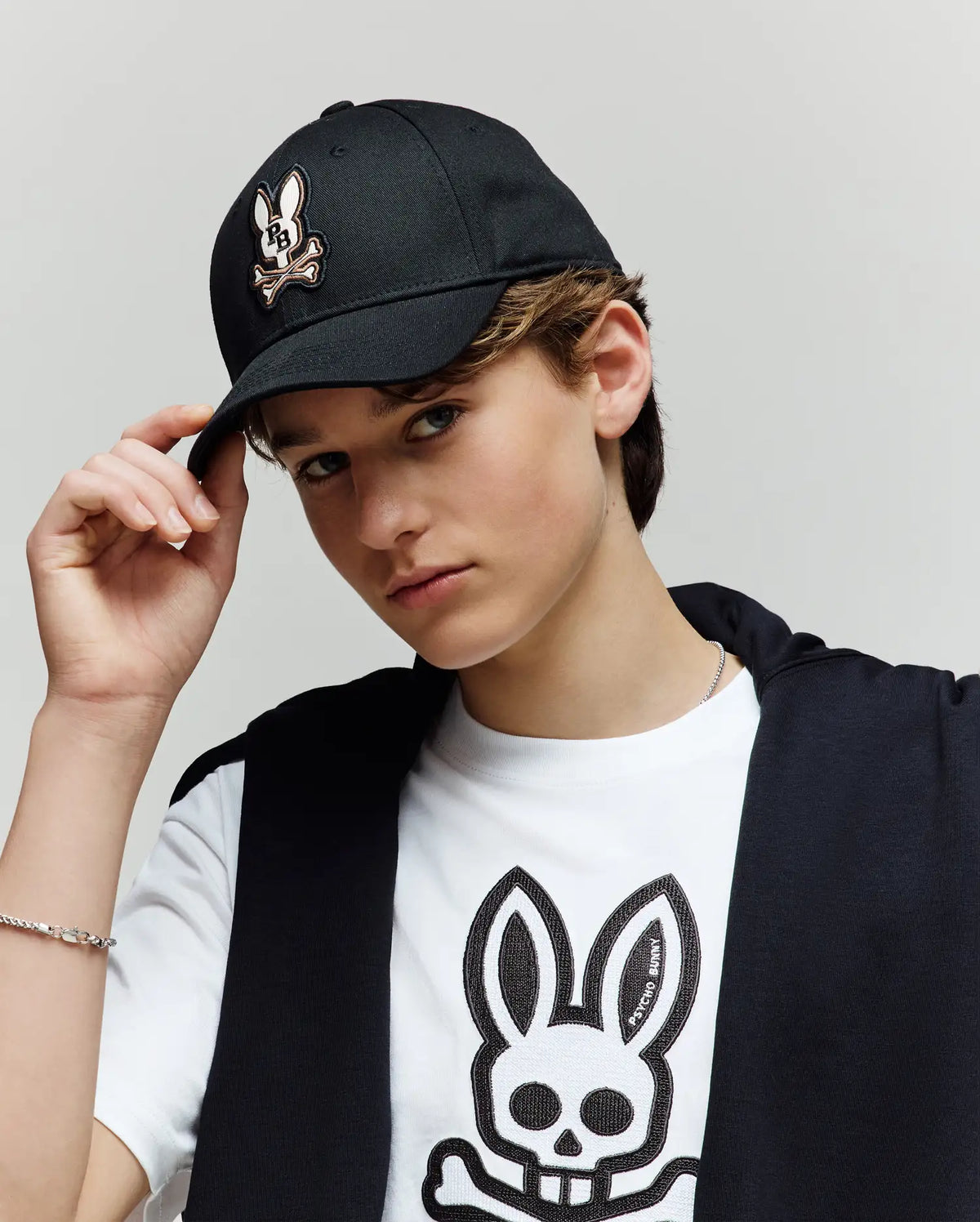 A young person wears the Psycho Bunny KIDS WADE BASEBALL CAP (KO1500016) with its embroidered Bunny skull logo, pairs it with a white tee and dark jacket, and looks at the camera while holding the cap's brim.