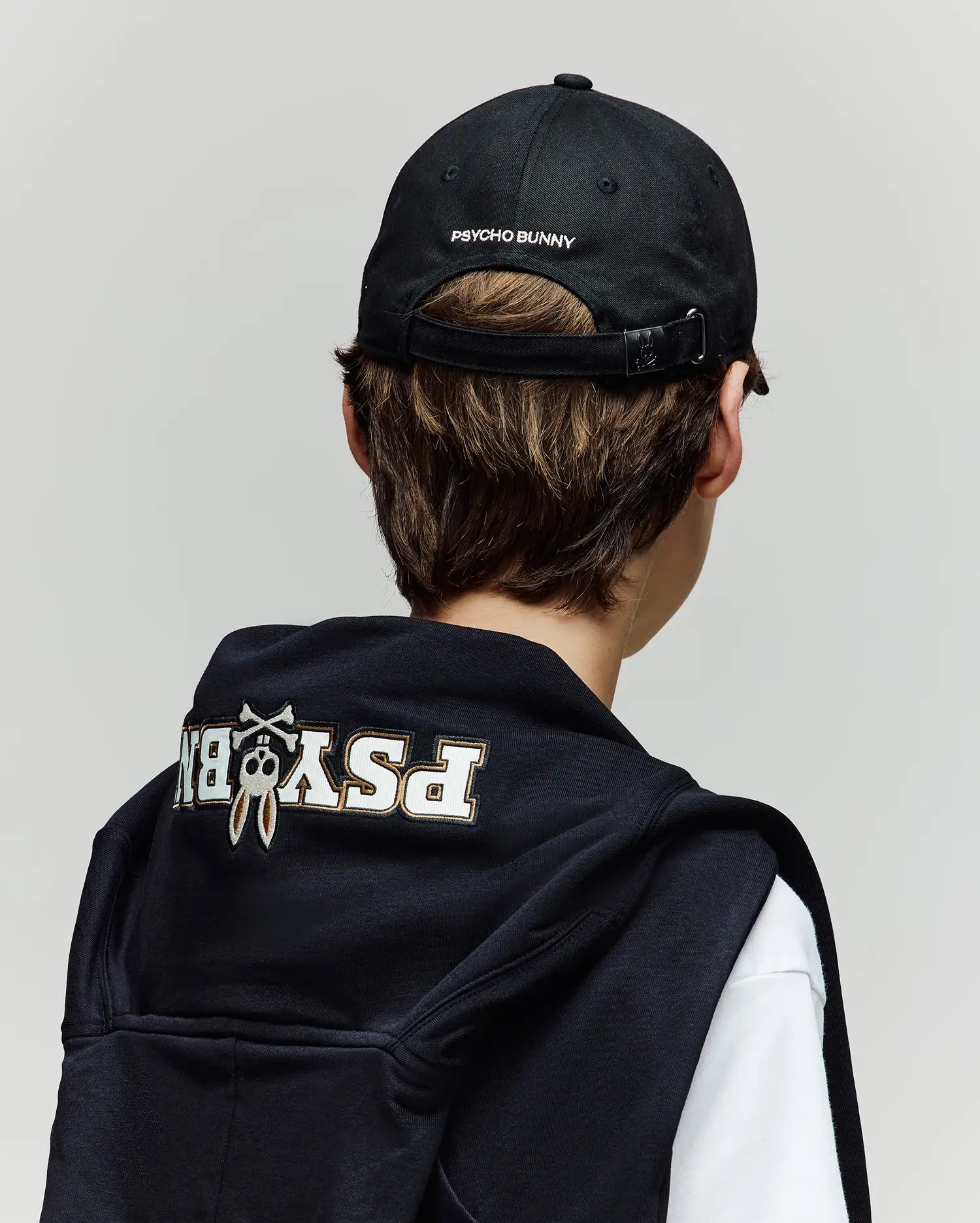 A person with brown hair is seen from behind wearing a Psycho Bunny KIDS WADE BASEBALL CAP (KO1500016) with an embroidered Bunny and logo, along with a black cotton twill hoodie draped over their shoulders.