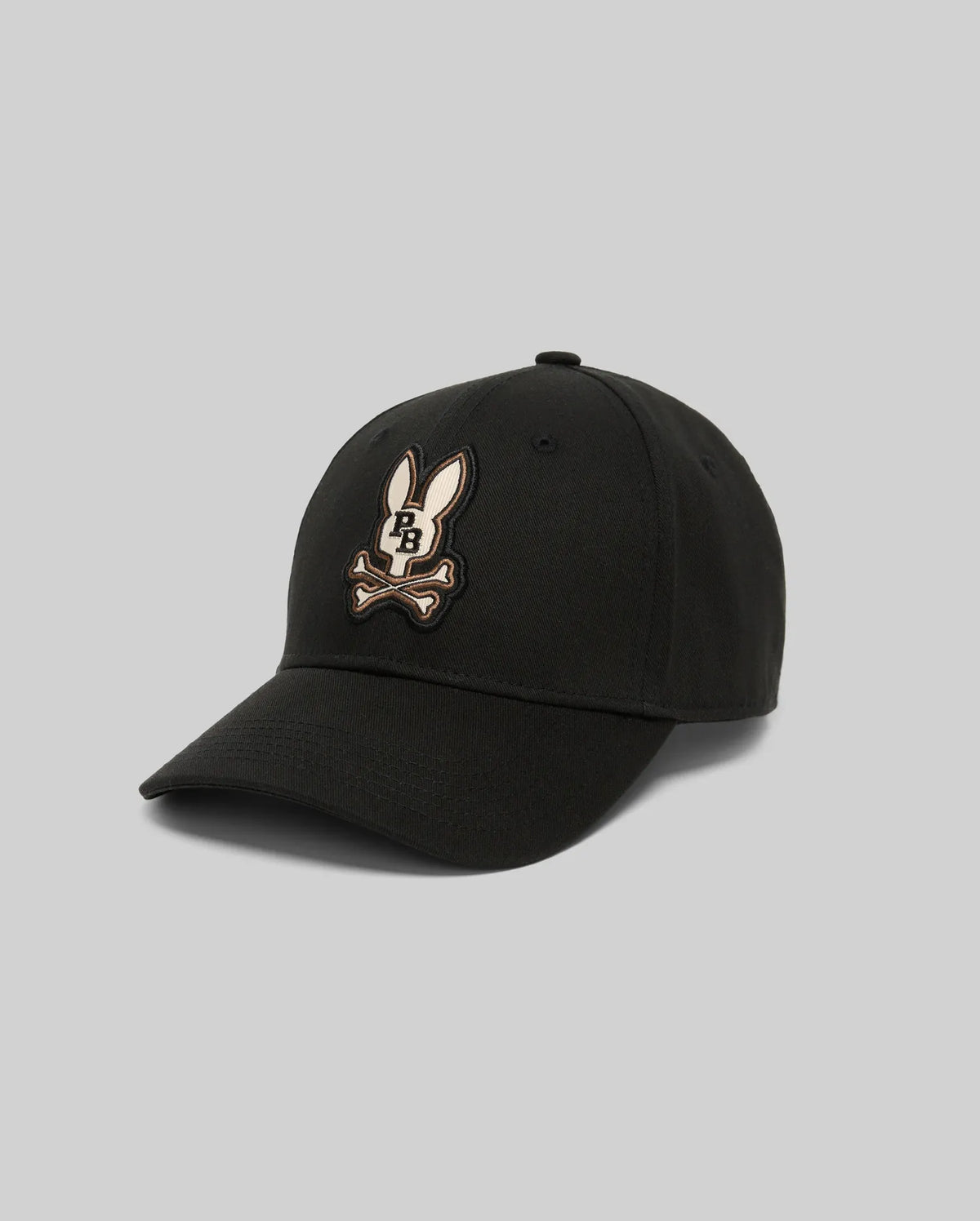 The KIDS WADE BASEBALL CAP - KO1500016 by Psycho Bunny is a black cotton twill cap featuring an embroidered cartoon bunny head over crossed bones on the front, shown against a plain gray background.