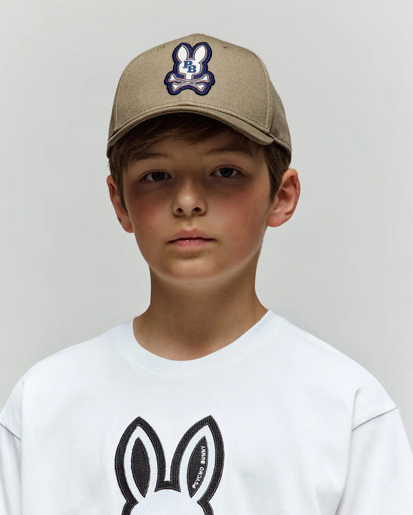 A young boy wears the Psycho Bunny KIDS WADE BASEBALL CAP - KO1500016 in tan cotton twill with an embroidered Bunny patch and a white tee featuring a black bunny face outline, standing against a plain light background.