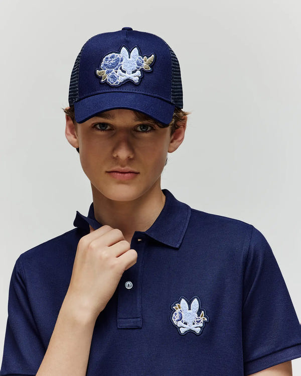 A young person wears the Psycho Bunny KIDS AUGUSTE TRUCKER CAP (KO1500022), a navy blue hat with an adjustable snapback and a white-blue chenille rabbit patch, paired with a matching polo, posing against a plain light background.