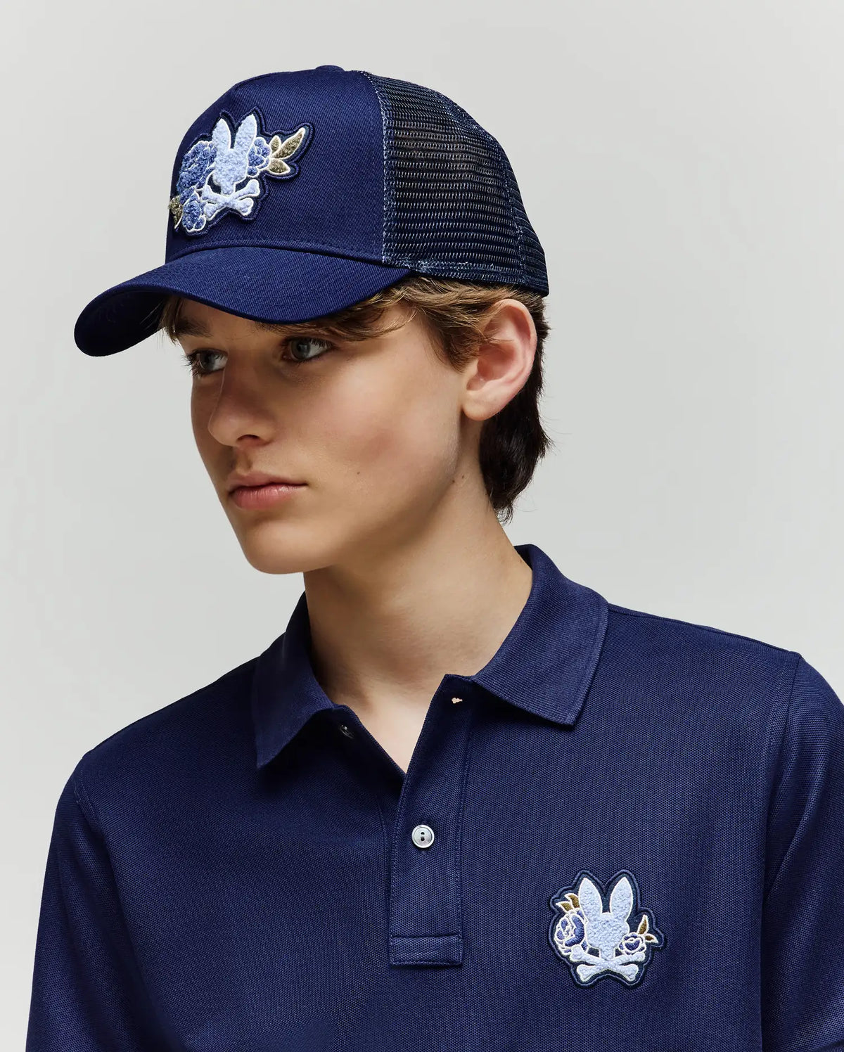 A young person wears the Psycho Bunny KIDS AUGUSTE TRUCKER CAP (KO1500022), a navy mesh hat with adjustable snapback and cartoon chenille patch, paired with a matching polo, both standing against a plain light background.