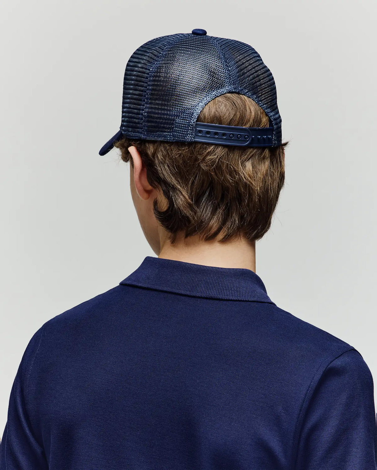 A child with brown hair is seen from behind, wearing the Psycho Bunny KIDS AUGUSTE TRUCKER CAP (KO1500022), a navy blue mesh snapback hat, and a matching navy blue collared shirt against a plain light background.