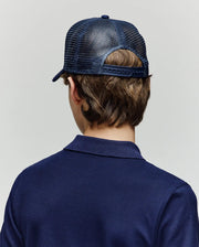 A child with brown hair is seen from behind, wearing the Psycho Bunny KIDS AUGUSTE TRUCKER CAP (KO1500022), a navy blue mesh snapback hat, and a matching navy blue collared shirt against a plain light background.