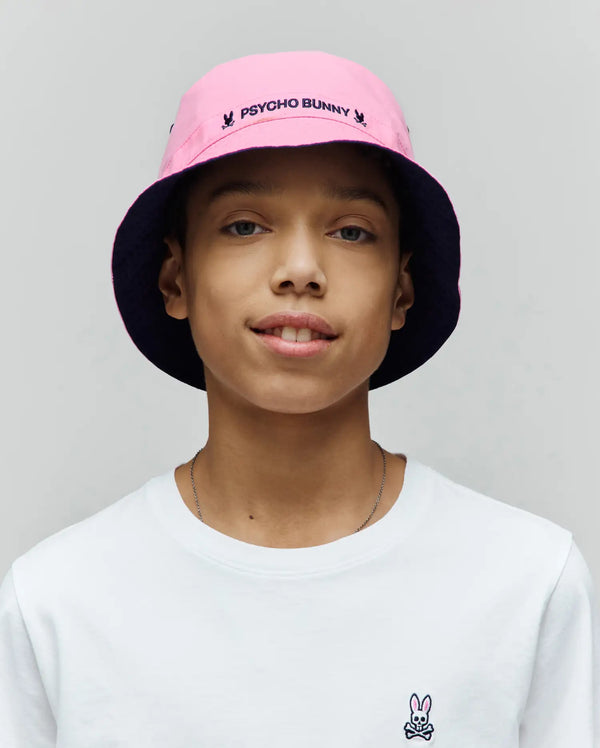 White classic fit jersey t-shirt with a small embroidered Psycho Bunny skull logo on the left chest, paired with a Kids Luke Reversible Bucket Hat in pink with a navy underside and embroidered Psycho Bunny text.