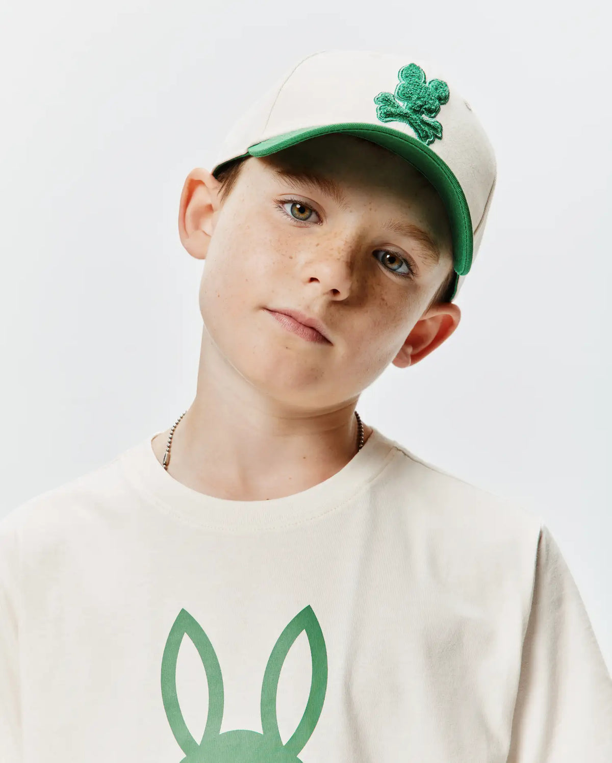 Beige KIDS CRAIG BASEBALL CAP with a green brim and embroidered Psycho Bunny skull logo, complementing an off-white jersey t-shirt featuring a large green bunny ears graphic on the chest. (1)