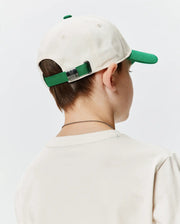Kids Craig Baseball Cap, an off-white cotton twill cap with a green brim and matching adjustable strap, featuring a silver Psycho Bunny skull logo buckle. Worn with a cream classic fit jersey t-shirt. (3)