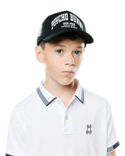 White classic fit pique polo with navy striped collar/cuffs and small embroidered Psycho Bunny skull logo, paired with a Black Kids Nathaniel A-Frame Trucker Cap featuring large white PSYCHO BUNNY text. (1)