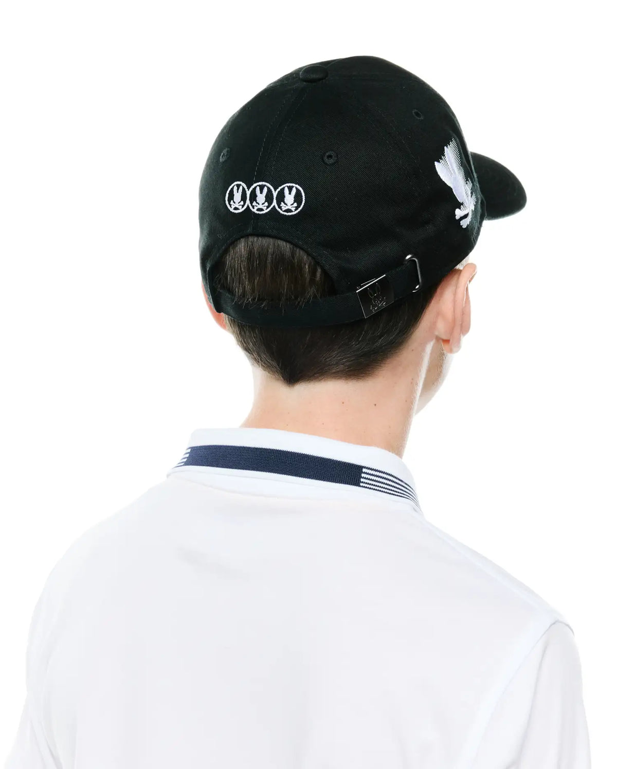 Black Kids Nathaniel A-Frame Trucker Cap with white embroidered Psycho Bunny skull logos on back and side, metal logo buckle, paired with a white polo featuring a navy and white striped collar. (3)