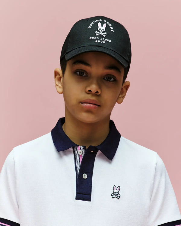 A boy in a white collared shirt with an embroidered Bunny logo and a Psycho Bunny KIDS ARCHIVE BASEBALL CAP (KO1500004) made of cotton twill stands against a pink background, looking directly at the camera with a neutral expression.