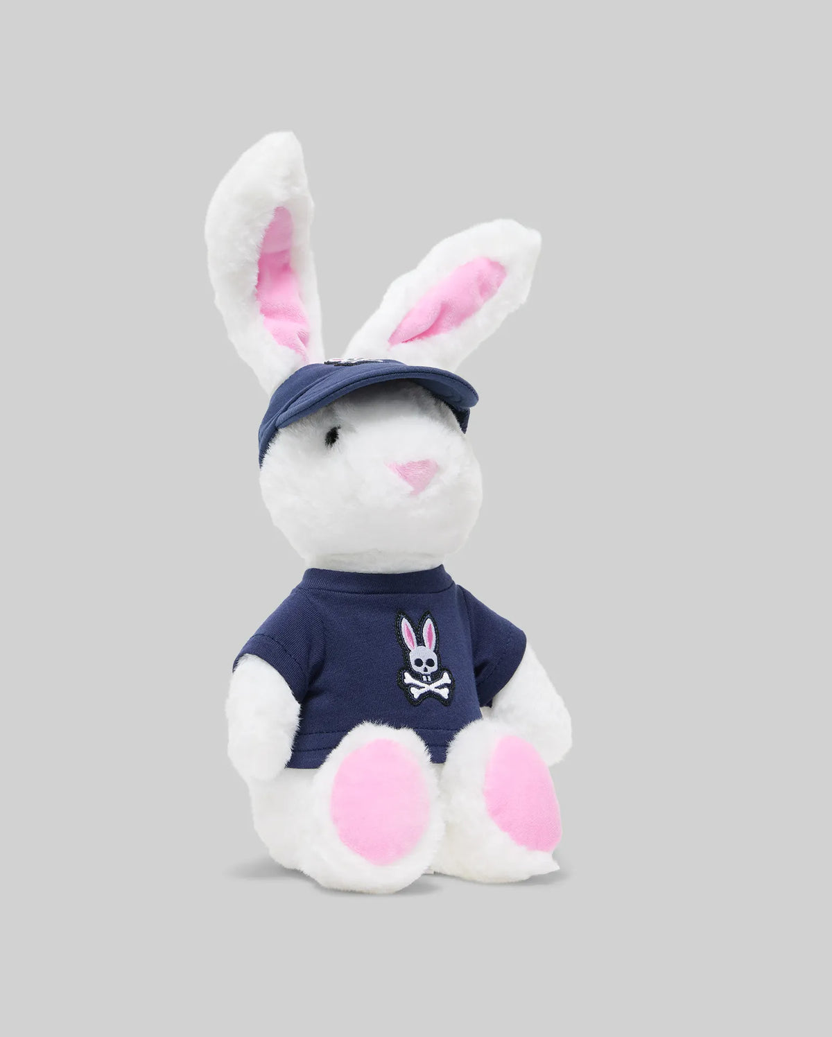 The PSYCHO BUNNY PLUSH - KO1700001 by Psycho Bunny features ultra-soft fabric, pink ears and feet, a navy blue cap and matching shirt with a bunny skull and crossbones logo—perfect for standing out against any light grey backdrop.