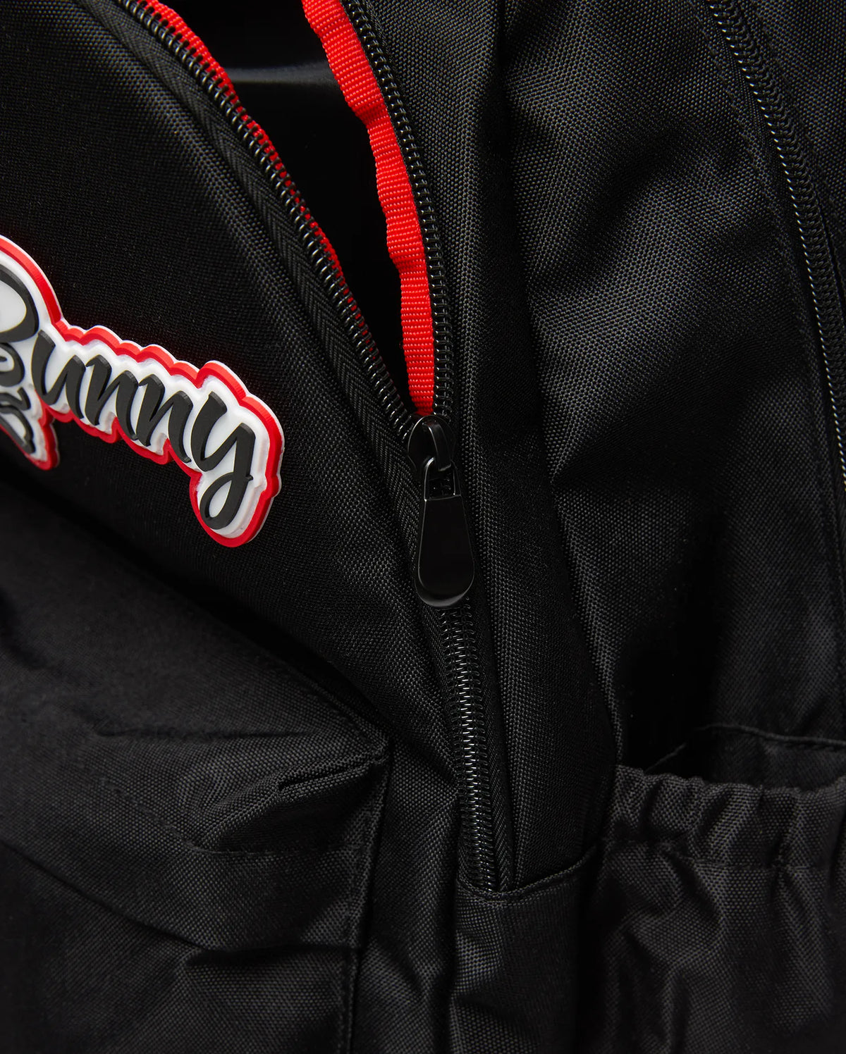 Black Kids Pal Backpack of durable woven fabric, featuring a raised black script Bunny logo with white and red outlines, a black zipper with red lining, and an elasticated side pocket. (3)