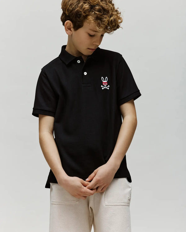 A young boy with curly hair looks down while wearing the Psycho Bunny KIDS MISCHIEF BUNNY PIQUE POLO (KT0100018), a black shirt featuring mother of pearl buttons and an embroidered bunny and crossbones logo, paired with light pants.
