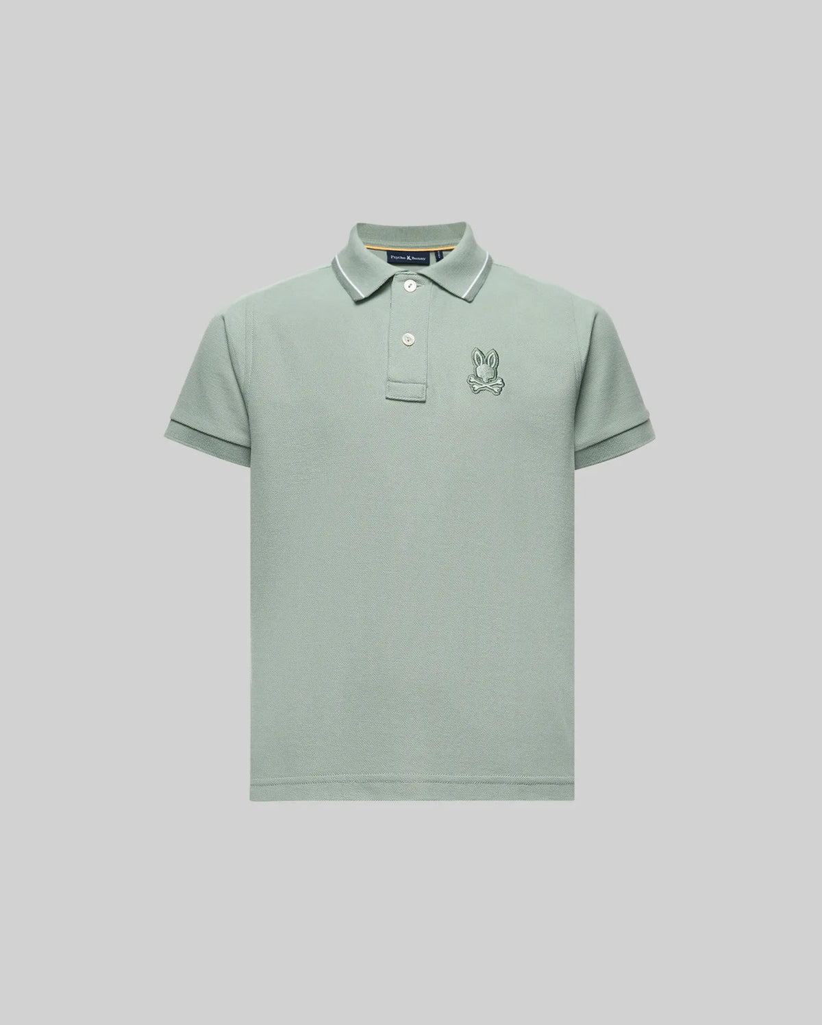 The Psycho Bunny KIDS FENLEY PIQUE POLO (KT0100023) features a light green color, regular fit, soft Pima cotton, short sleeves, collar, button placket, and an embroidered cartoon rabbit logo on the left chest. Shown on a light gray background.