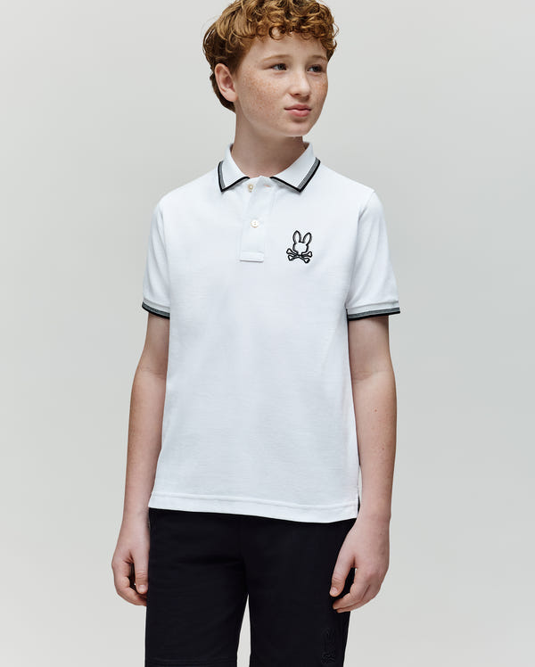 A boy with curly red hair wears the Psycho Bunny KIDS JUDE PIQUE POLO - KT0100027 in soft Pima cotton with an embroidered Bunny logo, paired with black shorts. He stands against a light gray background, looking slightly to the side and smiling.