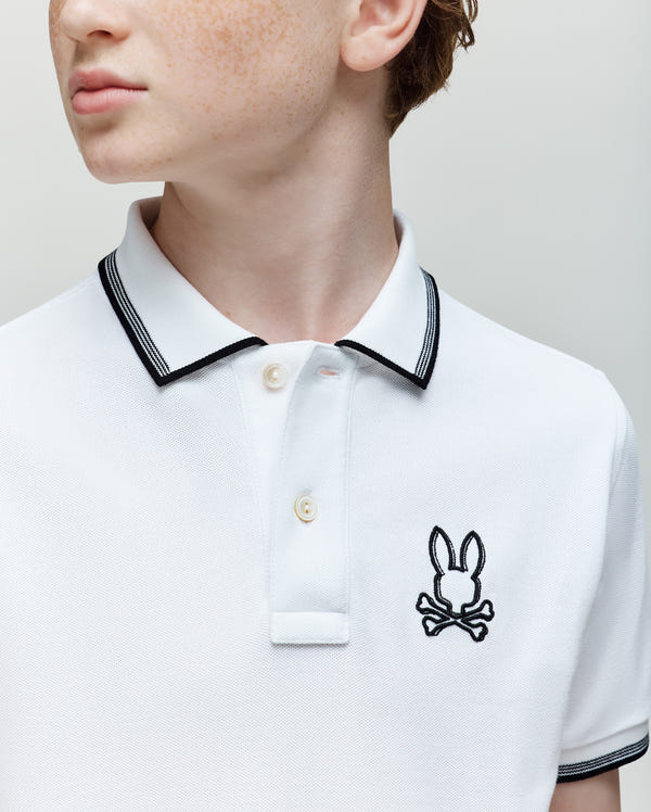 A young person wears the Psycho Bunny KIDS JUDE PIQUE POLO (KT0100027), a white Pima cotton shirt with black collar trim and an embroidered Bunny and crossbones logo, looking slightly to the side.