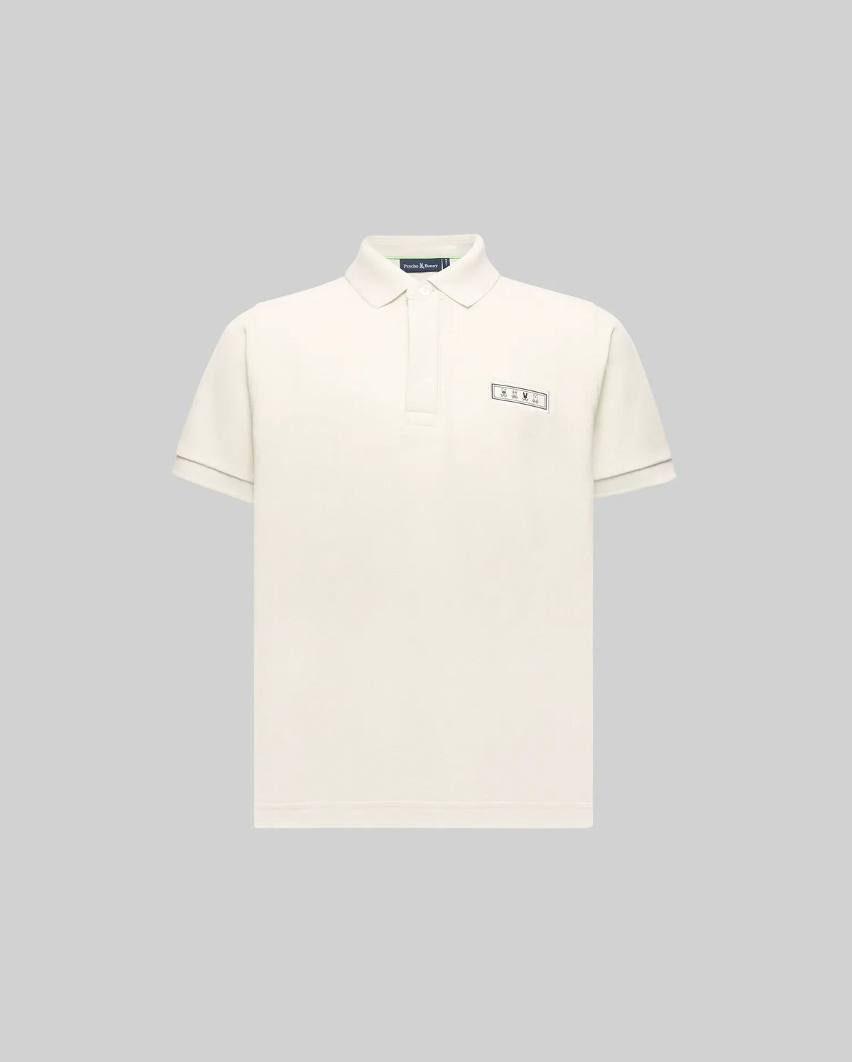 The KIDS AXTON PIQUE POLO - KT0100029 by Psycho Bunny is a plain, off-white, short-sleeved Pima cotton polo with a small rectangular jacquard-woven logo patch on the left chest, shown on a light gray background.