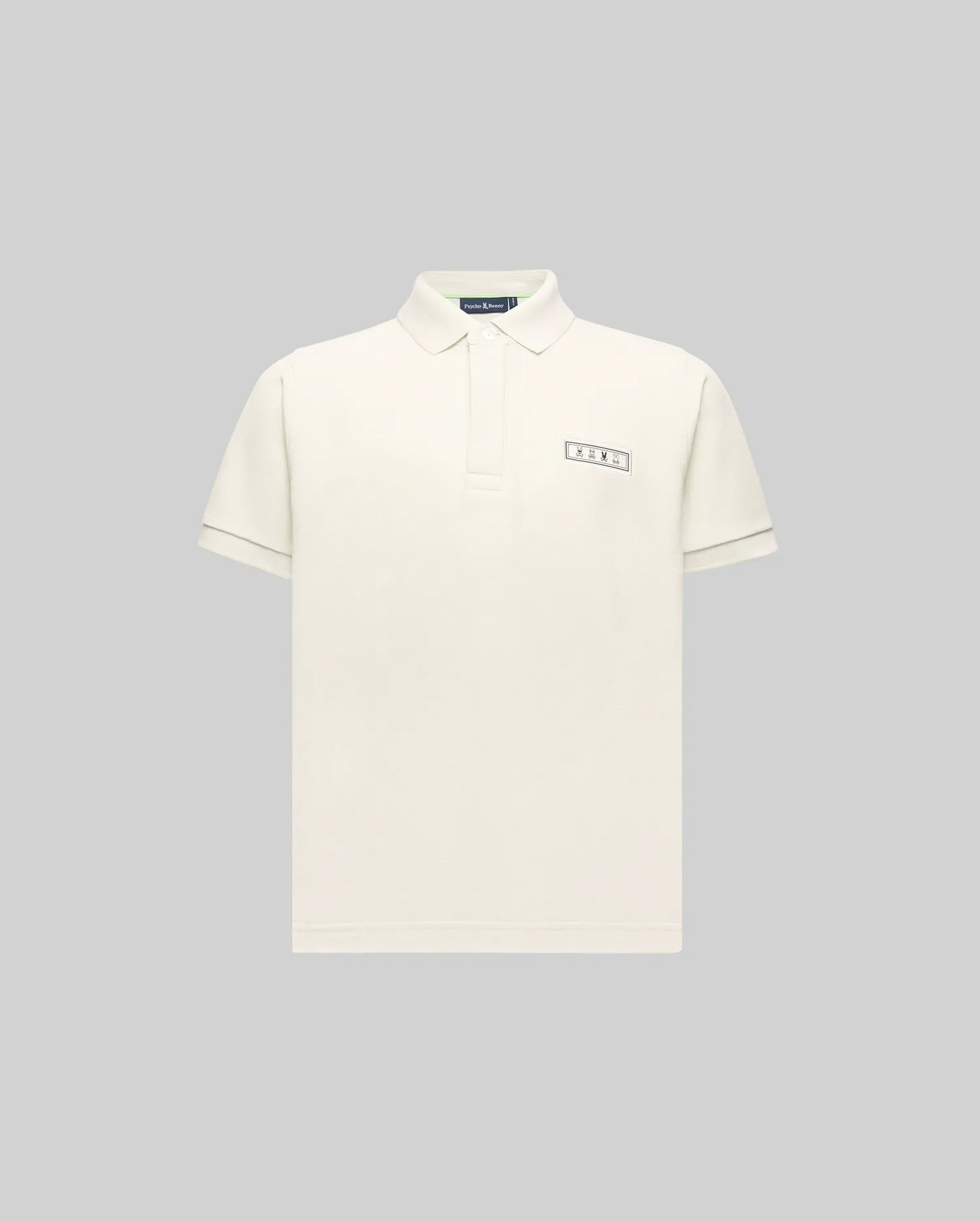 The KIDS AXTON PIQUE POLO - KT0100029 by Psycho Bunny is a plain, off-white, short-sleeved Pima cotton polo with a small rectangular jacquard-woven logo patch on the left chest, shown on a light gray background.