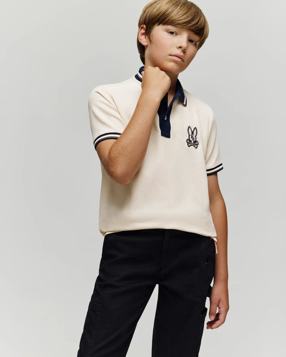A young boy with light brown hair poses confidently in a Psycho Bunny KIDS WADE PIQUE POLO SHIRT (KT0100032) with colorblock tipping and bunny logo, paired with black pants, against a plain light gray background.