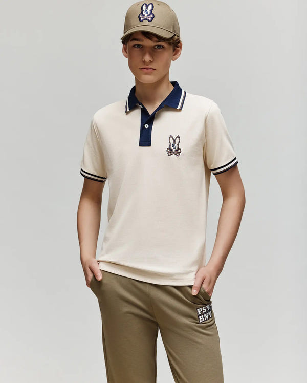 A young person stands with hands in pockets, wearing the Psycho Bunny KIDS WADE PIQUE POLO SHIRT - KT0100032 in beige with a cartoon rabbit patch, matching khaki pants, and a beige baseball cap with the same logo, against a plain gray background.